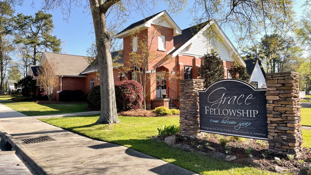 exterior photo of Grace Fellowship Church in Clanton, Alabama. Red brick building with Spring blooming trees and a wood and brick sign with the words Grace Fellowship "Come Discover Grace"